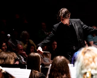 Conductor in black, with arms spread, amid heads of orchestra-members.