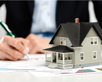 A miniature model of a house with a black roof and grey walls sits on a table with documents and cha