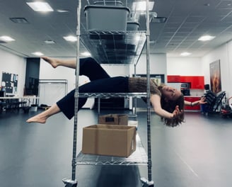 A young woman balances on a metal shelving unit, extending her limbs out and looking at the camera