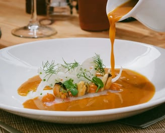 Sopa de pescado vertida en mesa con marisco, hinojo y hierbas frescas, junto a una copa de vino blanco