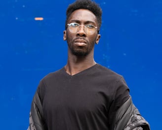 Man wearing circular wireframe glasses, black t-shirt and an Urban camo jacket.