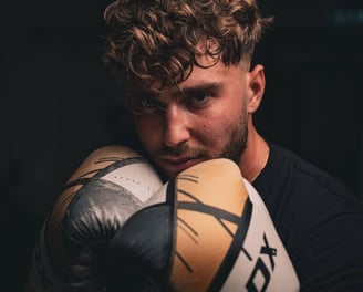 Barbat cu manusi de box RDX, in garda, priveste concentrat spre camera.