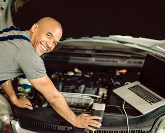 mecânico sorrindo trabalhando na parte elétrica do carro com um notebook aberto