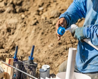 A plumber in a blue shirt is taping PVC piping on a construction site.