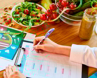 Mãos de nutricionista escrevendo em um prontuário sobre uma mesa com alimentos saudáveis