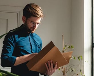 Bookkeeper standing up in a blue shirt looking at a client's folder to analyze their finances. A plant is to their left.