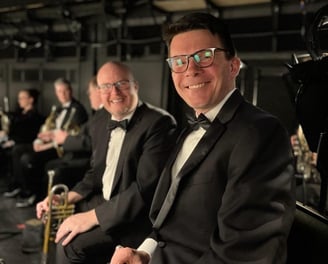 Two trumpet players wearing tuxedos, smiling in an orchestra pit.
