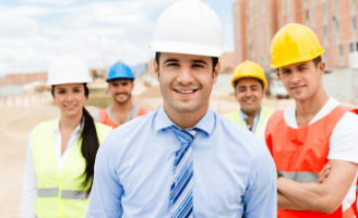 Engenheiro sorridente usando capacete branco e terno, com uma equipe de trabalhadores da construção civil ao fundo em um cant