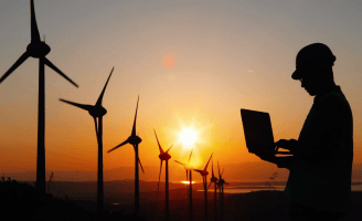 Engenheiro com capacete analisando dados em um laptop ao pôr do sol, com turbinas eólicas ao fundo