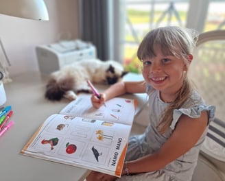 Mergaitė atlieka užduotis pratybose, kol katinas miega šalia