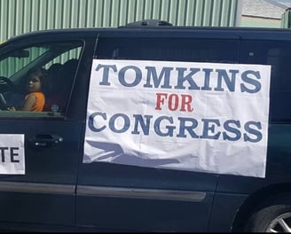 Banner with Tomkins for Congress on the side of a green minivan. A child can be seen in the window.