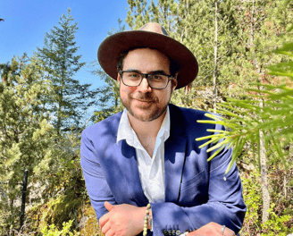 a smiling man in a suit and hat sitting on a bench in a forest