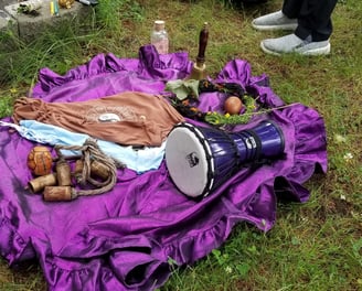Outdoor ceremonial setup on a purple cloth with a drum, bells, moon water, zodiac cloth, and ritual