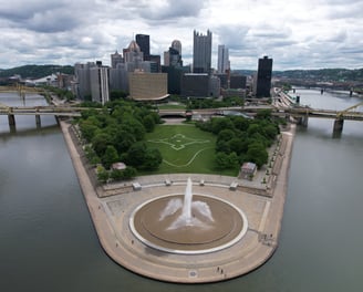 An aerial view of the Pitts Point Fountain in Pittsburgh, captured by a drone. The fountain sprays w