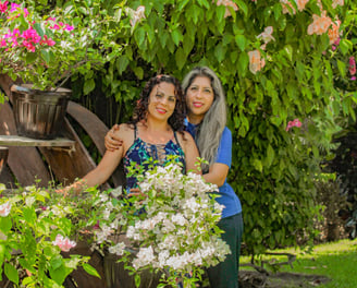 Fundadoras de Alianzas Ancestrales de pie en un jardín lleno de flores, una abrazando a la otra, reflejando unión, amor y con