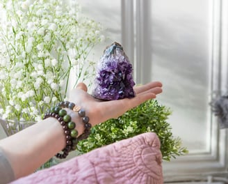 A photo of a hand holding a purple Amethyst cluster crystal with some white flowers