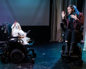 A white woman of short stature and a blue-haired person signing BSL, both in electric wheelchairs