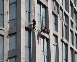 A person wearing a blue uniform and gloves is pushing a cleaning cart adorned with multiple small Union Jack flags. The environment appears to be a busy place, possibly a station or airport, with people in the background walking or standing.