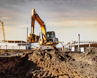 Bagger hebt Erde auf einer Baustelle aus bei Sonnenuntergang, im Hintergrund Rohbau und Kranstruktur