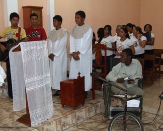 Pessoas celebram São João na igreja, com trajes religiosos e fiel em cadeira de rodas