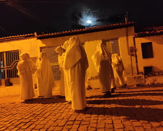 Penitentes cobertos de branco caminham à noite sob a luz da lua em rua de paralelepípedo