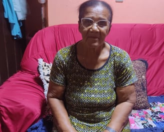 Senhora sentada em sofá vermelho, com roupas tradicionais, em casa simples e acolhedora