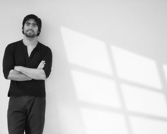 Hombre de cabello oscuro y barba, vestido de negro, apoyado contra una pared blanca con sombras geométricas de una ventana pr