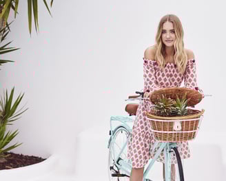 Jeune femme en robe à motifs assise sur un vélo Reid turquoise avec panier en osier contenant des pl