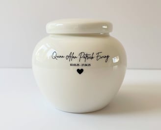 This is a rounded ivory ceramic urn engraved with the text Quinn Alan Patrick Ewing, and adorned with a small heart icon