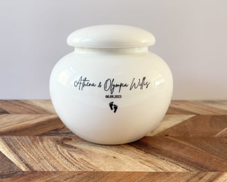 This is a rounded ivory ceramic urn engraved with the text Athena & Olympia Willis, and adorned with a small footprint icon