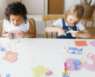 Two young girls cutting paper with scissors whilst making crafts at a table.