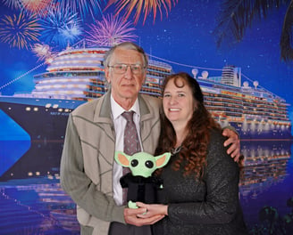 Man and Woman standing in front of a Cruise Ship holding a green stuffed animal between them.