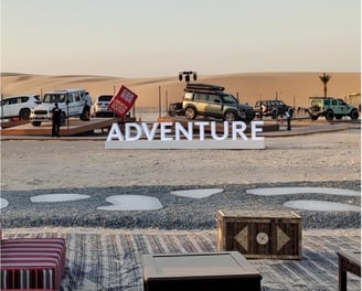 An outdoor car show setup in the desert featuring SUVs, a stage, and a large "ADVENTURE" sign.