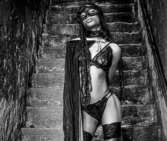 Photo artistique noire et blanc d'une jeune femme noire en sous vêtements et bas qui porte un masque dans le sous sol 