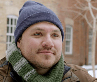 A young man in a scarf and toque smiles softly 