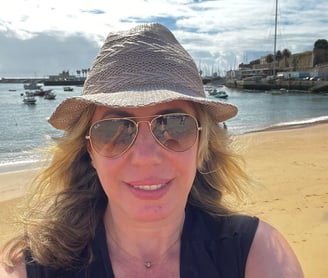 autora com chapéu e óculos de sol em praia com barcos ao fundo