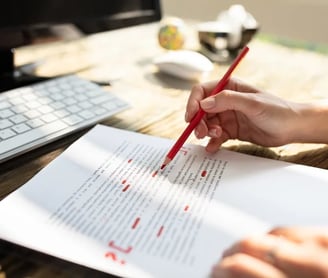 a person proofreading and correcting a document with a red pencil, computer in the background