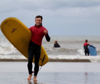 A stoked surf instructor carrying a yellow surfboard, giving a shaka and sticking his tounge out.