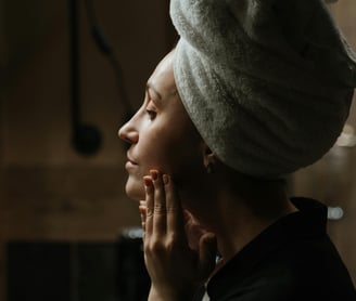 A woman in a robe studying her face in the mirror while her hair is wrapped in a towel.