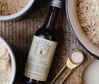 A bottle of vanilla extract beside bowls of rolled oats, brown sugar, and dough