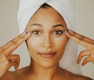 Woman with towel-covered hair applying a skincare product around her eyes