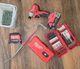 Milwaukee tools laid out on a hardwood floor with a box of nails