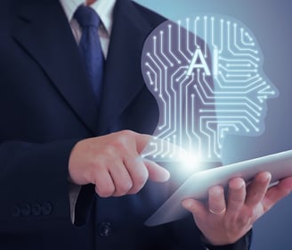 A business leader in a suit works on his tablet computer, with a holographic AI head emerging from the screen, face unseen.