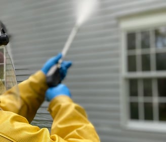 hands in blue plastic gloves hold a pressure washing lance pointing at a house wall in background