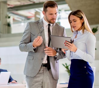 A female leader reviews data on a tablet with a male leader at the office, while their team works in the background.