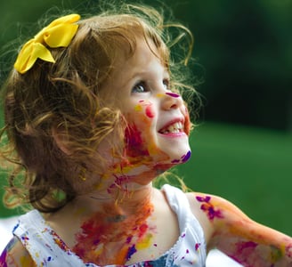 Petite fille souriante, cheveux en désordres, noeud jaune, pleine de peinture- Pourquoi micro creche