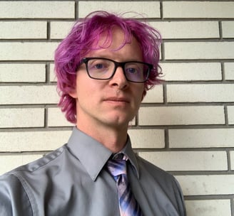 Tomkins with a grey shirt and blue and pink tie, with maroon colored hair and glasses.