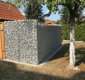 Eine als Sichtschutz zum Nachbarn errichtete Gabionenwand aus hellem Naturstein neben einem Holztor.