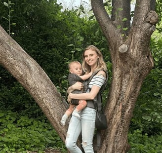 Mother holding baby in a carrier by a tree, bonding in nature.