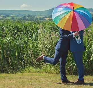 a gay couple on the day of the civil partnership kissing under a rainbow umbrella.
